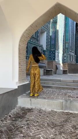 A woman in a yellow patterned outfit stands with her back to the camera, facing a large, ornate building with intricate turquoise, blue, and purple tiling. She is standing on a set of steps. To the right, another person wearing a pink and purple patterned dress and a headscarf enters the frame, holding a broom. The second person walks across the steps, sweeping the area. The first woman remains stationary, facing the building. The ground is paved with bricks. The scene appears to be in an outdoor courtyard.