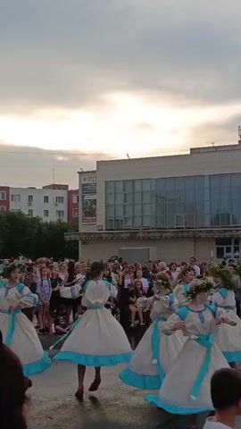 In this video, a group of young girls in traditional white and blue dresses with floral wreaths on their heads are dancing in a circle. They are outdoors, in what appears to be a town square or open public space. A crowd of people are gathered around, watching the performance. Behind the dancers and the crowd, there are buildings, including a multi-story residential building and a larger, modern building with a glass facade. The sky is overcast, suggesting a cloudy day, and the lighting indicates it could be late afternoon or early evening. The mood seems festive and lively, with the dancers appearing joyful and engaged in their performance. The location is identified as Krivodanovka, Russia. No specific text is clearly visible or legible in the frame.