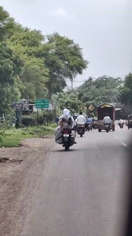 A motorcycle with two people riding it is traveling on a paved road. The rider and passenger are both dressed in traditional Indian attire. The passenger is wearing a white headscarf. The motorcycle is loaded with what appears to be luggage. Several other vehicles, including motorcycles, a truck, and a small pickup truck, are also visible on the road. The environment is rural, with trees and vegetation lining the sides of the road. The sky is cloudy.