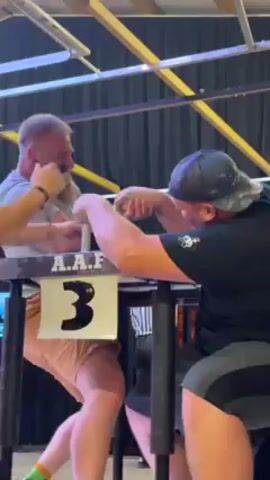 Here's a detailed description of the video:

The video shows two men arm wrestling at a competition. A blonde-haired woman in a striped referee shirt is standing beside the table, observing the match. The man on the left has a beard, short blonde hair, and is wearing a grey shirt. He has tattoos on his left forearm and is grimacing with effort. The man on the right is wearing a black t-shirt and a backwards baseball cap. He is also straining, but seems to have the upper hand. A number "3" is displayed on the table.

The arm-wrestling table is in the center of the frame. The table is dark, with a textured surface. The competition appears to be taking place indoors, under bright fluorescent lighting, with wooden rafters visible in the ceiling. There are curtains behind the table.

As the man on the left is defeated, the referee in stripes raises the arm of the man on the right in victory. Both competitors shake hands.