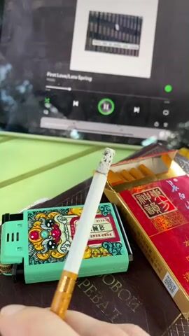 This media file captures a close-up, still life arrangement, focusing on smoking paraphernalia and a tablet device. The primary subjects are a lit cigarette held between fingers, a decorative lighter, and an open pack of red and gold branded cigarettes. In the background, a tablet screen displays what appears to be a music streaming interface, showing album art, song title "First Love/Late Spring" by "Mizuki", and playback controls.

The scene is set on a dark-colored, textured surface, possibly a book or a table, with a hint of a green surface in the upper left corner, suggesting an outdoor or well-lit indoor setting. The lighting suggests it could be daytime, with natural light illuminating the cigarette and casting subtle shadows. The cigarette is actively burning, with smoke rising from the tip, indicating the immediate moment of smoking.

The lighter is a prominent object, featuring intricate turquoise, gold, and red decorative elements, possibly in a traditional Chinese style. The cigarette pack is also vividly colored in red and gold, bearing Chinese characters and what looks like a QR code. The visible text on the cigarette pack includes Chinese characters and "0.5 mg". The text on the lighter appears to be Chinese characters as well.

The overall mood evoked is one of personal relaxation and indulgence, with the music playing on the tablet adding to the ambiance. The focus on the cigarette and lighter suggests a deliberate composition, possibly capturing a moment of quiet contemplation or a break. The specific mention of Dujiangyan City, China, provides a geographical context, hinting at the cultural origins of the cigarette branding and possibly the design of the lighter.