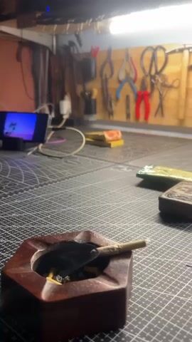 The video shows a close-up of a wooden ashtray with a lit cigarette in it. Smoke is rising from the cigarette. In the background, a workbench is visible with various tools hanging on the wall, including pliers and scissors. There is also a smartphone displaying a colorful image, and a few boxes on the workbench. A person's arm with tattoos reaches into the frame, seemingly to adjust something out of view. The lighting suggests an indoor setting, possibly a workshop or studio.