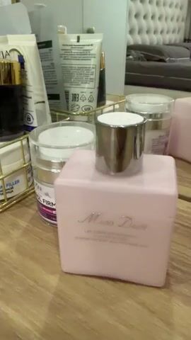 The video shows a close-up, stationary shot of several cosmetic bottles and jars arranged on a wooden surface, likely a vanity or dresser. In the foreground, a pink rectangular bottle labeled "Miss Dior" is prominent, with a silver cap. To its left, a tall, frosted green bottle with a light green spherical cap is partially visible, with text that appears to read "PISTACHIO CRUSH" and "KOTINNT". Behind these, several other cosmetic containers are visible, including tubes and jars with gold and white accents. The background is out of focus, but suggests a bedroom setting with a headboard and a mirror reflecting some of the items. The lighting is soft, highlighting the products. The overall impression is one of personal grooming or skincare items.