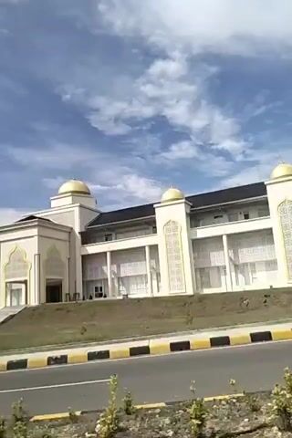 A video pans across a modern, cream-colored building with gold domes and intricate architectural details reminiscent of Islamic design. The building appears to be a large public structure, possibly a mosque or a government building, situated on a green lawn with a road in the foreground. The sky is blue with scattered white clouds, suggesting a clear, daytime setting. The camera movement is smooth, offering a consistent view of the building's facade. No people are visible in the frame.