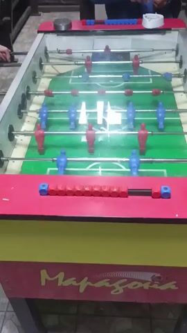 The camera is positioned to capture a foosball table, where two people on each side are playing. The table has a clear glass top and a green playing surface with blue and red figures. Red scoring markers are present at the bottom of the table. The table's design includes a red and yellow base with the name "Maradona" printed on it. The table is situated on a tiled floor.