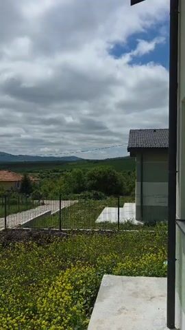The video shows a rural landscape. In the foreground, there are patches of yellow flowers and a concrete slab. A fence separates the foreground from a grassy field with scattered trees and houses. One house has a red tile roof. A gravel driveway leads towards the houses. In the background, there is a range of hills and mountains. The sky is overcast with a mix of dark and light clouds. Power lines are visible. The camera pans slightly to the left.