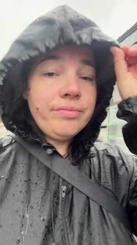 A woman in a black rain jacket with the hood up is speaking into the camera as rain falls, creating water droplets on her jacket. She has dark hair, light skin, and a small mole on her left cheek. Her right hand is near her head, adjusting her hood. The background shows blurred greenery and other people walking.