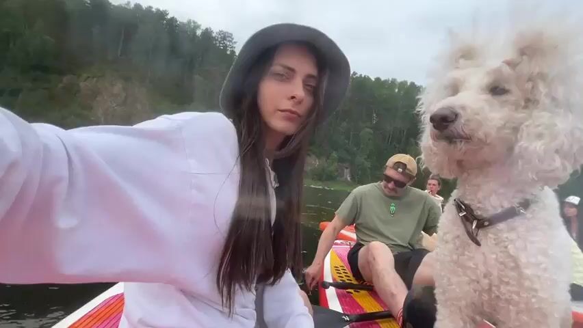 A group of people and a dog are on paddleboards in a river. A young woman in a white hoodie and green hat is in the foreground, taking a selfie with a white poodle standing next to her on the paddleboard. A man wearing sunglasses and a green t-shirt is sitting on another paddleboard behind them. In the background, there are trees and a rocky cliff. The water is calm, and the sky is cloudy.