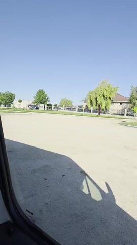 The camera is in a vehicle, looking out through the window. The view shows a bright blue sky above a large, mostly empty beige or light gray parking lot. In the distance, there is a house behind a black wrought-iron fence, with trees planted around the property. Further back, a body of water can be seen on the horizon. The shadow of a hand is visible on the car window. Music is playing in the background.