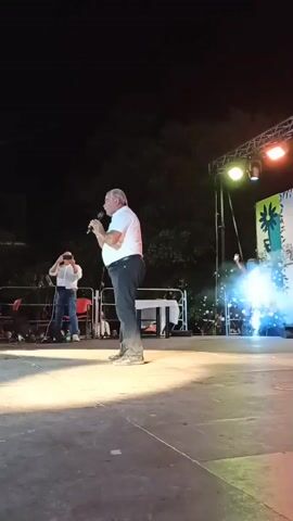 An outdoor nighttime performance is captured from a low angle, focusing on a man on a stage. He is wearing a white polo shirt, dark pants, and light-colored shoes, and is holding a microphone in his right hand. He appears to be singing or speaking into the microphone, with his left hand raised slightly. In the background, there is a stage with lighting equipment and a backdrop that is partially obscured. To the left of the main performer, another person is visible, appearing to hold up a phone, possibly recording. To the right of the main performer, a person in a red top is dancing or moving energetically, with smoke or fog effects creating a hazy atmosphere around them. The stage is illuminated by bright stage lights, casting a strong glow on the performer and the stage floor. The background is dark, indicating it is night.