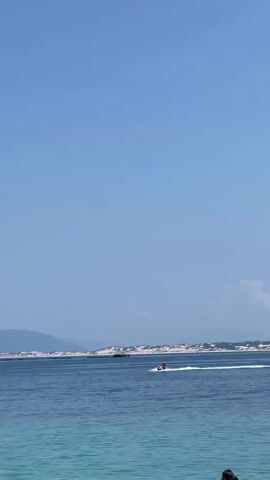 The video shows a bright, clear day with a vast expanse of blue sky. The ocean is calm, with a lighter blue color in the foreground that deepens to a darker blue further out. In the middle ground, a person is riding a red jet ski, leaving a white spray trail behind them as they move across the water. In the background, there is a sandy coastline with some vegetation and hills or mountains. A few scattered clouds are visible in the sky, and a white boat is docked on the right side of the frame. The scene is serene and evokes a sense of a typical summer day at the beach.