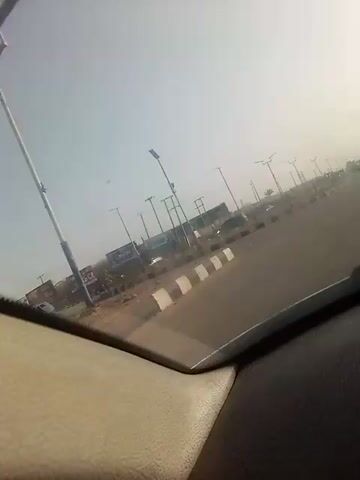 The video shows a view from inside a car moving along a road. The road is lined with streetlights and power poles. There are a few other vehicles on the road. The sky is hazy. Concrete dividers are visible. Buildings and signage are visible in the background. A pedestrian is crossing the road. The interior of the car is visible in the foreground.