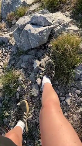 The video begins with a shot of a person's legs from their perspective standing on a rocky, grassy landscape. The person is wearing shorts, white socks, and black Adidas sneakers. The camera angle then elevates to reveal a panoramic view of a hilly, green landscape leading to a body of water. The sun is shining brightly, reflecting off the water, and the horizon is visible in the distance. The foreground consists of rocky terrain with patches of green vegetation, while the background features a vast expanse of green hills leading down to the water.