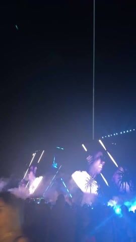 The video shows a concert at night. In the foreground, a crowd of people is visible, some dancing. Above the crowd is a stage with large screens displaying the face of a man and palm trees. Bright lights and lasers shine from the stage, creating a vibrant, energetic atmosphere with fog. A tall vertical light pole is visible in the background against the dark night sky.