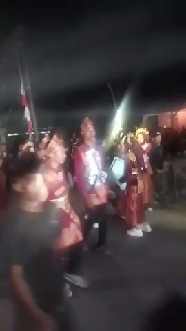 A group of people in traditional red and gold attire, possibly performing a cultural dance or parade, are captured in a low-light, outdoor setting at night. The individuals, primarily young men, wear conical hats and elaborate vests adorned with gold trim. They are in motion, some appearing to march or dance. A bright light source, possibly a torch or a camera flash, momentarily flares up in the foreground, obscuring some of the detail. In the background, indistinct figures and structures suggest a public gathering or street event. The overall atmosphere is festive and celebratory, despite the challenging lighting conditions.
