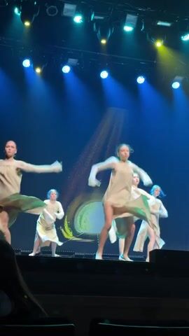 The video shows a group of dancers on a stage performing a routine. The stage has a dark blue backdrop with a large, glowing circular design in the center that appears to be part of the set, possibly a projection. There are bright blue and yellow stage lights illuminating the performers from above.

Five dancers are visible, all wearing light-colored, flowing dresses or tunics. They appear to be young women or teenagers.

At the beginning of the video, the dancers are spread across the stage. Two dancers on the right side of the frame are low to the ground, with their bodies extended horizontally, mimicking a prone position, then transitioning into a kneeling position. Another dancer on the far left of the frame is also in a low position, though less visible. Two other dancers are briefly seen in the background or moving into frame.

The dancers are moving fluidly and synchronously. The choreography involves dynamic transitions from standing to low positions on the floor, incorporating arm and leg extensions. The lighting changes slightly during the video, emphasizing certain areas of the stage. The focus of the camera is on the dancers and their movements. The perspective is from an audience member's view, slightly elevated and looking at the stage.