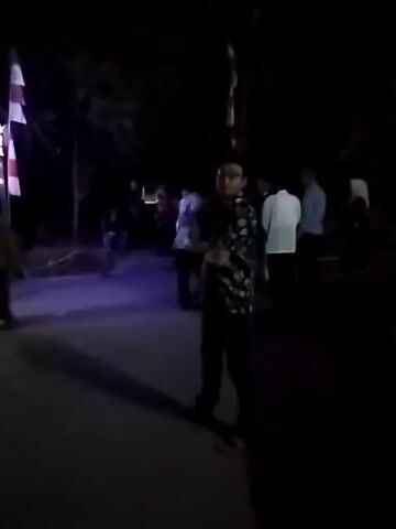 This is a low-light video of an outdoor nighttime scene, possibly a street or pathway.  A decorative archway adorned with white lights and red and white flags is visible on the left. Several people are present, some standing and conversing, while others are walking. The overall atmosphere appears to be a gathering or celebration.