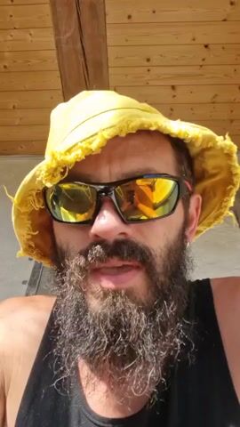 A man with a beard and a yellow hat is looking at the camera. He is wearing sunglasses and a black tank top. The background is a wooden ceiling. The video is a selfie taken by the man.