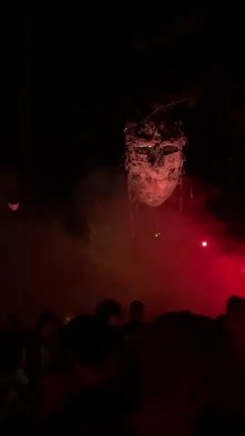 The video shows a nighttime outdoor gathering in a wooded area. A large, abstract structure resembling a face or mask hangs suspended from trees. The scene is illuminated by colored lights, predominantly green and red, which create dramatic lighting effects and cast shadows. Smoke or fog machines are in use, adding to the atmospheric lighting. A crowd of people, silhouetted against the lights, are gathered below, facing the illuminated structure. The overall impression is that of an art installation or performance event in a natural setting at night.