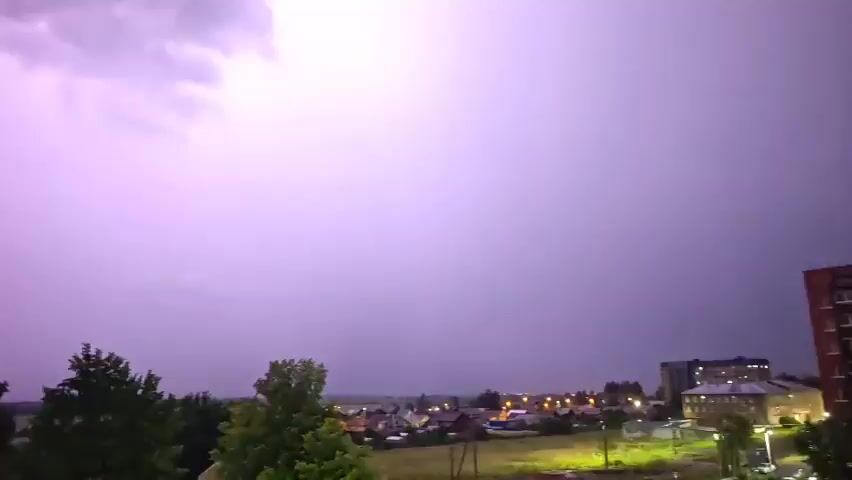 The video shows a night sky over a town. A distant lightning flash briefly illuminates the dark clouds and the buildings below. The lightning is faint and appears as a white streak against the black sky. The town lights are visible as a warm glow on the horizon. There are trees in the foreground, their silhouettes dark against the sky. The video is dark and grainy, suggesting it was taken with a low-light camera.