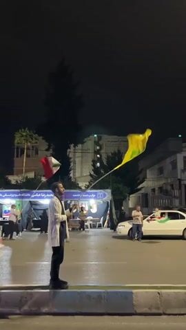 The video shows a man in a white coat standing on a street at night, holding a yellow flag with a green emblem. He is in the foreground and appears to be addressing someone or participating in an event. In the background, there are tents set up on the side of the road, with people milling around. There are also cars driving by, and buildings in the distance. Another person is visible in the background, holding a different flag. The overall atmosphere seems to be a public gathering or demonstration.