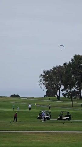 The video shows a golf course on an overcast day. Several golfers are scattered across the green, some are preparing to swing, one is in mid-swing, and others are walking. Golf carts are parked on the course. In the sky, a paraglider can be seen in the distance. The environment appears to be coastal, with a glimpse of the ocean visible in the background. The overall mood is relaxed, typical of a golf outing.