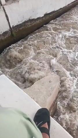 The video shows a powerful, turbulent flow of water, likely in a river or a large canal, in Mandihar, Pakistan. The water is murky brown with significant white frothing and eddies, indicating a rapid current.

In the foreground, a concrete barrier or railing is visible, with a person's foot in a black flip-flop briefly seen at the beginning, suggesting the viewer is standing on or next to this structure. A concrete block is also visible near the water's edge, partially submerged in the churning flow.

As the camera pans, a wider view reveals the full extent of the waterway. A long, white concrete fence or railing runs along one bank, separating the water from an area with trees and vegetation. The water body extends into the distance, flanked by green banks and trees under what appears to be an overcast or hazy sky. The consistent design of the waterway and the structures suggest it is an engineered canal system, possibly experiencing high flow due to heavy rainfall or water release. The activity is purely the dynamic movement of the water. No text is visible.