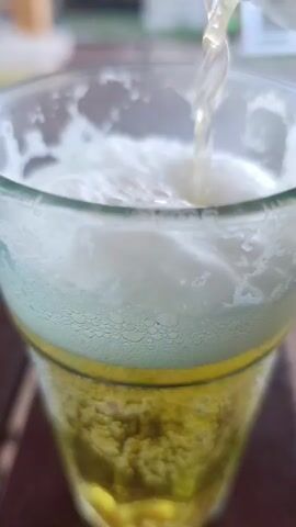 The video shows a close-up shot of beer being poured into a glass. The beer is a light golden color and is frothing as it is poured. The glass is clear and has a yellow band around the top. The background is blurred, but appears to be an outdoor setting with wooden furniture. The video is very short, only a few seconds long.