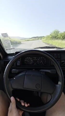 A POV shot from inside a car shows a person's hands on the steering wheel as the car drives down a rural road. The dashboard and windshield are visible. The road is surrounded by grassy fields and trees, with hills in the distance under a clear blue sky. The sun is shining brightly.