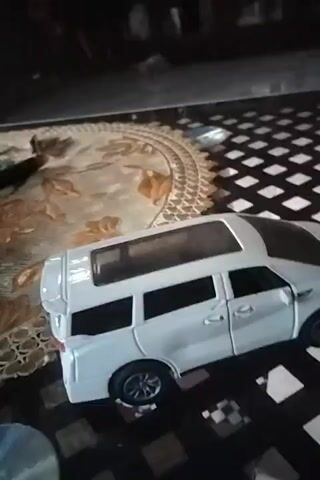 The video shows a white toy van with a black roof on a black and white checkered surface. To the left of the van is a round floral tablecloth. The background is dark.