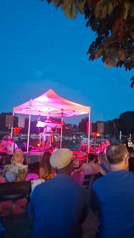 This media file captures an outdoor music performance at dusk in Wiesbaden, Germany. A band is set up under a large, white, diamond-shaped tent that is illuminated with colorful LED lights, transitioning through pink, blue, and green. The band members are visible, with at least one person playing an instrument, likely a keyboard or synthesizer, and another singing.

Several people are gathered around the stage, observing the performance. Some are seated at tables in the foreground, while others stand. The atmosphere appears relaxed and festive, suggesting a casual concert or event. The sky is a deep blue, indicating that it is evening. Trees and foliage are visible in the background, framing the scene. In the distance, lights suggest the presence of buildings or other structures, possibly near a body of water given the reflections. The overall mood is pleasant, with the vibrant lighting adding to the celebratory feel of the gathering. No discernible text is visible in the frame.