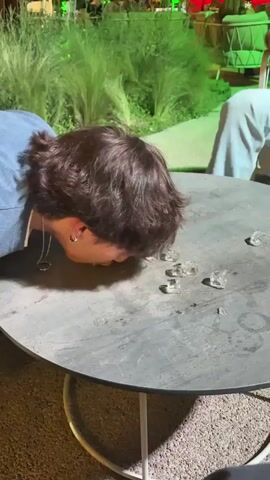 The video captures a playful evening scene at an outdoor bar or restaurant patio, likely in Turin, Italy. Two individuals are featured around a round, dark grey table with a white metal base. The scene is illuminated by ambient artificial lights, including vibrant green and subtle red accents, indicating it's nighttime.

The central activity involves a person with dark, medium-length hair, wearing a blue top, leaning down with their face close to the table. They appear to be attempting to pick up scattered ice cubes from the table surface using only their mouth, suggesting a game or challenge. Several clear ice cubes are visible on the wet surface of the table.

A second person, wearing a brown jacket and light-colored pants, is partially visible. They are seen placing or picking up a tall glass containing a yellowish drink, observing the activity with amusement. Another similar drink glass rests on the table.

The background features lush green foliage, suggesting a garden or well-landscaped area, with other blurry patrons and outdoor seating visible. The ground beneath the table is a light-colored gravel. The atmosphere is lighthearted and social, underscored by laughter audible in the background, implying enjoyment and amusement from the participants and onlookers. There is no visible text in the image.