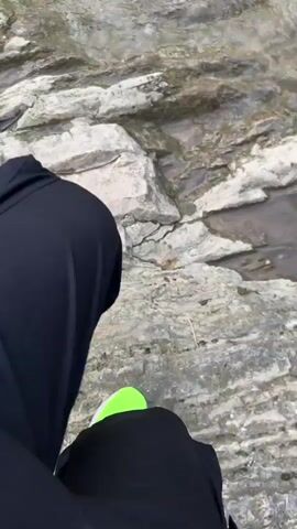 The video starts with a close-up of a person's legs and bright green sneakers as they stand on a rocky outcropping near a body of water. The camera then pans up to reveal a scenic waterfall surrounded by rocky cliffs and sparse greenery. The waterfall feeds into a pool of turbulent water. The sky is overcast.