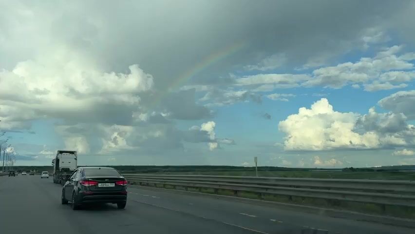 This is a wide-angle, first-person perspective video from a vehicle traveling on a highway. The camera faces forward and slightly to the right, capturing the road ahead and the surrounding landscape. Several cars and a large truck are visible on the highway, moving in the same direction as the camera. The sky is a mix of dramatic clouds, with some dark, heavy clouds on the left and brighter, more scattered clouds on the right. A faint rainbow is visible arcing across the sky. The road is asphalt with clear lane markings. A metal guardrail runs along the right side of the highway. Beyond the highway, there is a flat, green landscape, possibly fields or marshland, stretching to the horizon. The overall impression is of a journey on a highway under a dynamic sky with a hint of post-rain weather.