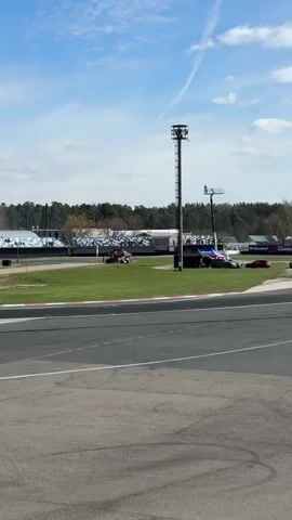 The video shows two cars drifting around a corner on a racetrack. The first car is a black and yellow BMW, while the second car is a dark blue sports car. Both cars are leaving trails of white smoke behind them as they drift. The racetrack is surrounded by grass and trees, and there are bleachers in the background with spectators. The sky is blue with some clouds.