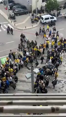 From an elevated angle, the video shows a large crowd of people gathered on a tiled square adjacent to a street intersection. Many are wearing yellow and black clothing, suggesting they might be supporters of a sports team. People are milling about, and some are moving in different directions, occasionally bumping into each other. A few individuals seem to be jogging or running within the crowd. A red barrier is partially visible at the edge of the intersection. The street features parked cars and motorcycles, and a white van is also present. A street light pole is situated close to the perspective of the camera. The video appears to capture a moment of activity or unrest amongst the crowd.