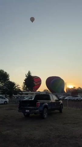The scene shows an outdoor setting at dawn or dusk. The sky is a light blue, and the sun is near the horizon, creating a warm, golden glow. Three hot air balloons are visible in the sky. One is already airborne and relatively small, while the other two are on the ground, still being inflated. In the foreground, there's a dark-colored pickup truck parked on a grassy or dirt area. A fence runs along the background, separating the area with the balloons from a parking lot filled with cars. A few trees and a palm tree are also visible, contributing to the scenery. The ground appears somewhat uneven, possibly indicating a field or open space. The overall atmosphere is peaceful and serene, with the focus on the preparation of the hot air balloons against the backdrop of the rising or setting sun.