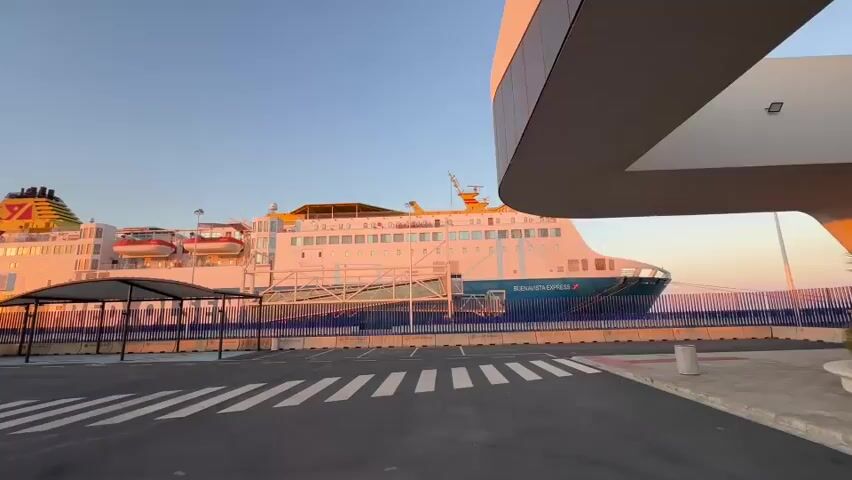 The video shows a large ferry docked at a port. The sun is low in the sky, casting a warm glow on the ship and the surrounding area, suggesting either sunrise or sunset. The ferry, named "Buenavista Express," is white with blue and orange accents. It appears to be a passenger ferry, with multiple decks and lifeboats visible. In the foreground, there is a parking lot with marked spaces and several cars parked under shaded shelters. The ground is asphalt, with white lines indicating parking spots and pedestrian crossings. A modern-looking building with a curved roof is partially visible on the right side of the frame. The sky is clear and a soft gradient of blue and orange. There are no people visible in the video. The overall scene is calm and serene, highlighting the juxtaposition of the large vessel and the empty port facilities.