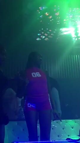 A group of people are dancing in a dimly lit club with colorful lights. Two women in the foreground are dancing together, and one is wearing a pink tank top with the number "06" on it. A DJ booth with blue lights is visible in the foreground. The atmosphere is energetic and festive.
