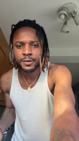 Here's a description of the video:
The video shows a man in a white tank top with dreadlocks standing in what appears to be a kitchen. He has a nose piercing and a gold necklace. He is holding a metal strainer or sieve over a white bowl. He dumps the contents of the strainer into the bowl, then shakes the strainer to try to dislodge any remaining substance. The strainer seems to have a handle on the side. The kitchen setting is somewhat visible, with a light fixture on the ceiling and cabinetry in the background. The man appears to be focused on the task of emptying the strainer into the bowl.