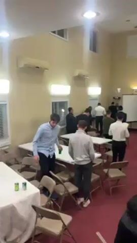 A mostly unoccupied room, furnished with white tables and tan folding chairs on a red carpet, is populated by around fifteen people. Most of them are male and appear to be young adults, and most of them are standing or walking. They are generally dressed in casual attire, including dress shirts and slacks. Several people are wearing yarmulkes. The room is brightly lit by overhead lamps. There is a pull-down projection screen at the back of the room, and air conditioning units are mounted on the walls.