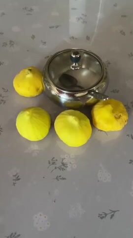 The video shows a close-up, slightly shaky, overhead shot of five yellow figs arranged around a metal sugar bowl with a glass lid and a spoon inside. The figs are plump and appear to be fresh, with some slight indentations on one of them. The sugar bowl is shiny and reflects the surrounding environment. The surface the objects are on is a light-colored tablecloth with a subtle floral pattern. The lighting is soft and appears to be coming from above, casting faint shadows. There are no people visible in the video, and no actions are being performed other than the slight camera movement. The scene is static and focused on the arrangement of the figs and the sugar bowl.