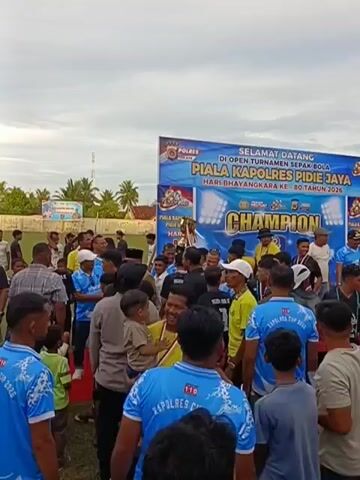 The video shows a gathering of people outdoors, possibly at a sporting event. A large banner in the background reads "SELAMAT DATANG DI OPEN TURNAMEN SEPAK BOLA PIALA KAPOLRES PIDIE JAYA BHAYANGKARA KE- 80 TAHUN 2023". Many people are wearing blue shirts with the text "KAPOLRES CUP 2023" and some wear yellow shirts. Several individuals appear to be children, one of whom is being held by an adult. Some people in the crowd are wearing police uniforms. The overall atmosphere seems festive and celebratory.