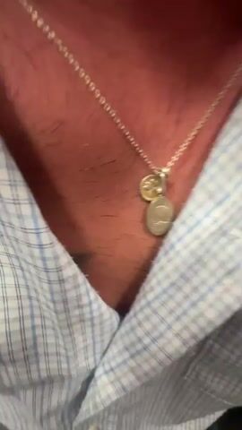 The video opens with a close-up shot of a person's neck and chest area, showing a silver chain with two pendants. The pendants appear to be a small round medallion and a larger oval-shaped one. The person is wearing a light blue and white checkered shirt, and some chest hair is visible. The background is out of focus. The second part of the video shows a selfie-style shot of a man's face. He is wearing the same necklace and shirt. His eyes are closed, and his tongue is sticking out. He appears to be in a public transportation setting, possibly a train, as a metal pole and another person are visible in the background. Sunglasses are perched on top of his head.