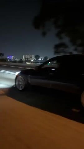 A dark-colored car with silver wheels drives on a highway at night. The car emits flames from its exhaust. The car passes under a highway sign, and the buildings of a city skyline can be seen in the distance.