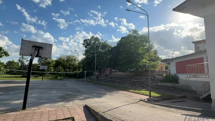 The camera shows an outdoor basketball and volleyball court, likely in a public park or schoolyard. The court surface appears to be concrete with faded painted lines. A basketball hoop stands prominently in the foreground, and a volleyball net stretches across the court. Tall streetlights are visible around the court. In the background, there are trees and some buildings, one of which has a set of concrete bleachers nearby. The sky is blue with scattered clouds, and the sun is shining. The scene appears empty and still, with no visible people or active play.