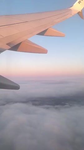 The video shows a view from an airplane window. The airplane wing is visible in the foreground, with the engine protruding from the bottom. Below the wing, a thick layer of clouds stretches out to the horizon. The sky above is a gradient of blue, with soft pink and orange hues near the horizon, suggesting either sunrise or sunset. The overall scene is serene and vast, emphasizing the height and distance from the ground.