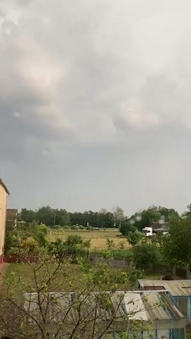 This media file captures a landscape scene from a window, possibly in the Матыкальскі сельскі Савет area of Belarus. The dominant feature is the sky, filled with heavy, grey, and dark storm clouds, indicating overcast and potentially rainy weather. The overall mood is somber and anticipatory, suggesting an approaching or ongoing storm.

Below the sky, the scene transitions to a rural or suburban environment. A building, partially visible on the left, appears to be a house or apartment block. In the middle ground, there is a field or open grassy area, bordered by trees and a fence. Further into the distance, more trees and scattered houses can be seen, including what looks like a white van or truck parked near a building on the right.

In the foreground, branches of a leafy tree obscure parts of the view, suggesting the camera is positioned indoors looking out through a window. On the lower right, parts of a greenhouse with a translucent roof and a light blue frame are visible, further contributing to the impression of a residential, perhaps rural or semi-rural, setting.

The presence of water droplets on the windowpane is a notable detail, reinforcing the idea that it is raining or has recently rained. The time of day is difficult to ascertain precisely due to the cloudy conditions, but the lighting suggests it is daytime. There are no people clearly visible in the frame. No text is visible in the media file.