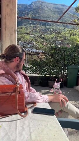 The video shows a man sitting at a table on a patio, with a phone and bag beside him. He has long hair tied back, glasses, a striped shirt, and a sweater draped over his shoulders.  The camera pans to the right, revealing a large, white, fluffy dog yawning on the ground. Behind the dog are a green trash bin, a stack of red bricks, and a washing machine. A toy stroller is visible in the background. The patio is surrounded by greenery, and a mountain is visible in the distance under a clear, sunny sky.