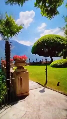 The video shows a picturesque garden setting on a bright, sunny day. A paved walkway leads the eye across a lush green lawn. A sculpted tree with a rounded crown is prominent in the garden. A decorative lamppost stands near the tree. In the background, figures of people are visible along a stone railing, with a lake and mountains further in the distance. A tall stone planter filled with red flowers is in the foreground. A palm tree is partially visible on the left side of the frame. Bird song can be heard.